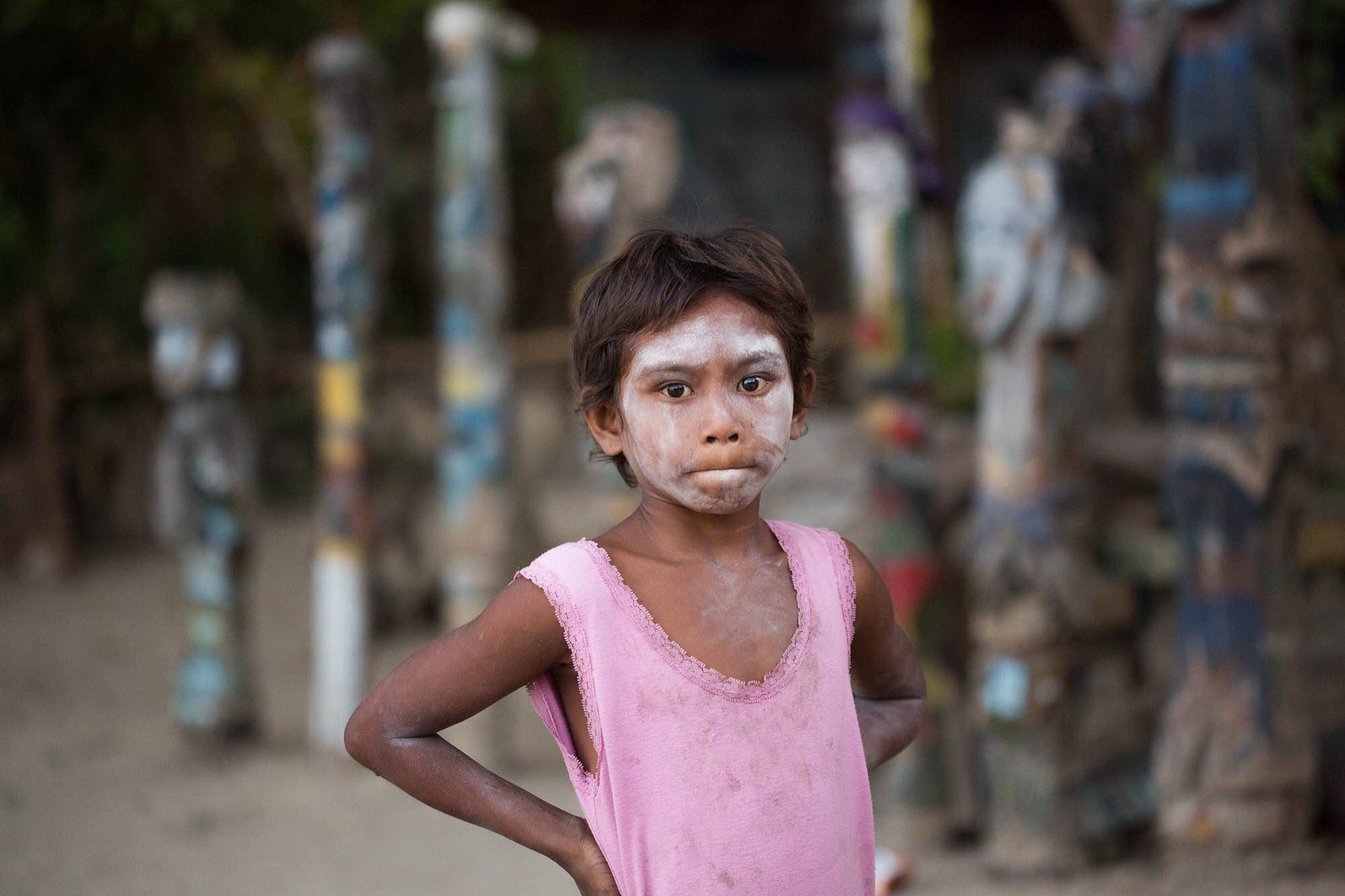 A young Moken boy with his face covered in Thanaka takes a break from playing games in front of the village's spirit poles.